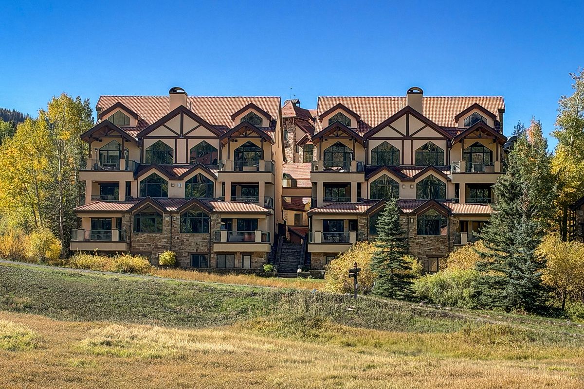 Dakota Place in early October: two matching stucco, timber, and stone buildings under tile roofs, with golden aspens and a blue sky above the ski run