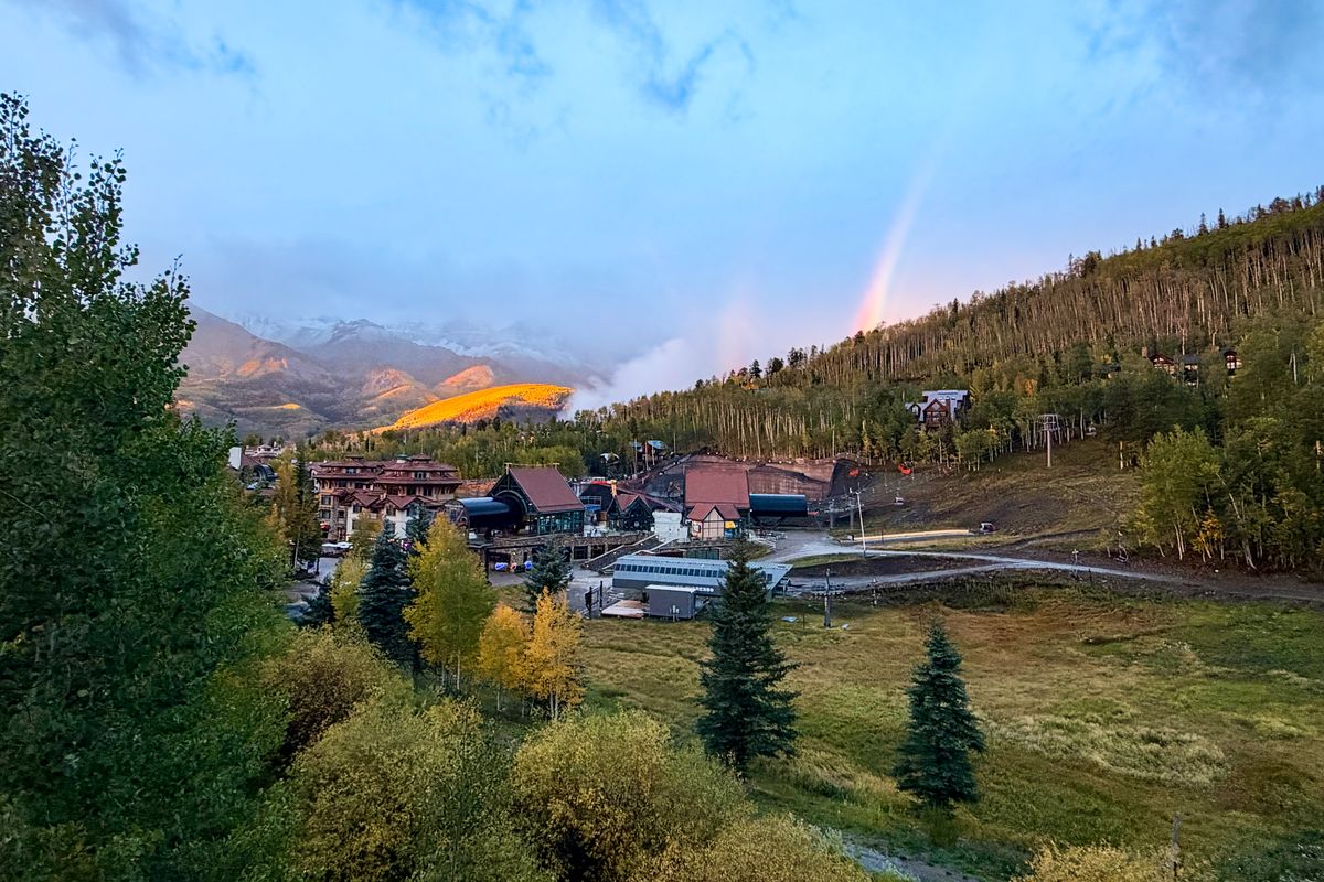 View from a third-floor balcony at Dakota Place over the Mountain Village core: the gondola station and Village Express lift below, golden aspens on the slopes, and a rainbow over the San Juan Mountains